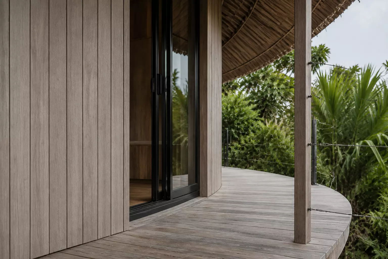 Terraza curva de madera que rodea un bungalow, entre la pared de madera y la vegetación.