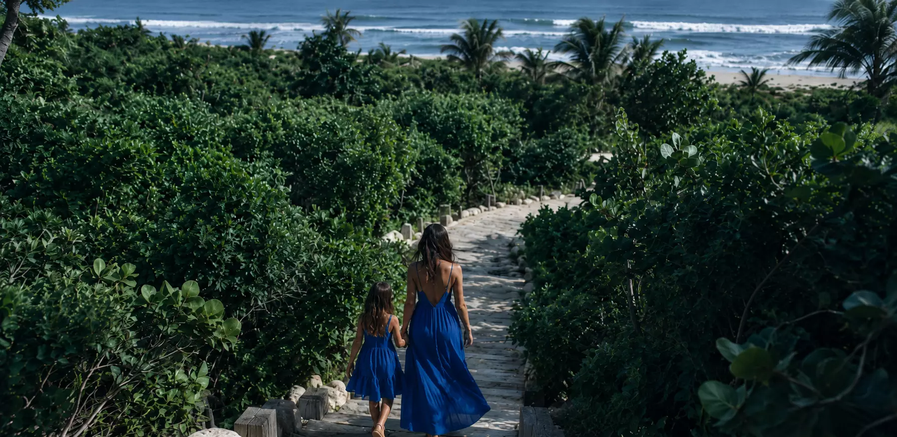Una mujer y una niña bajan por un camino de piedra entre la vegetación hacia la playa.